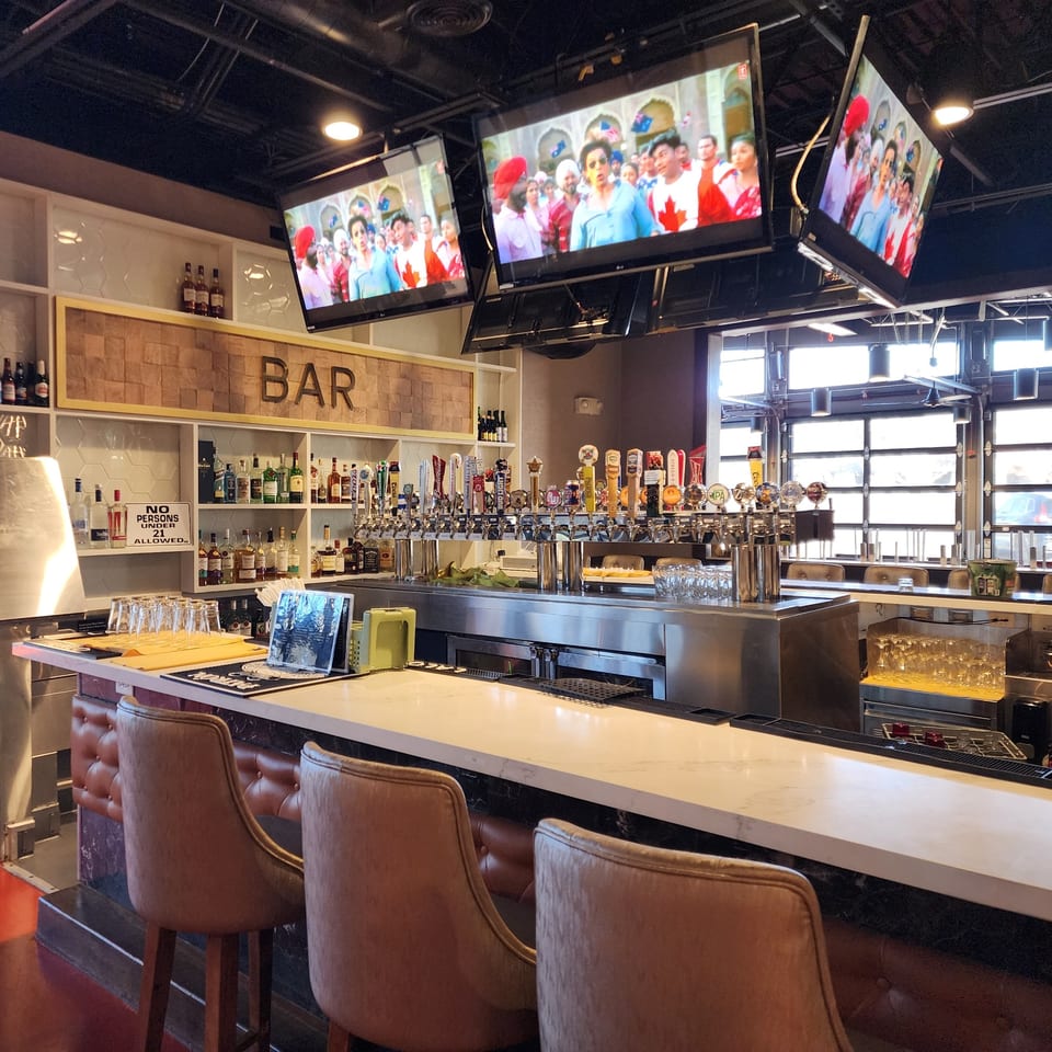 A well-stocked bar at Garnish Restaurants with multiple beer taps, liquor bottles, and TVs showing Indian content.