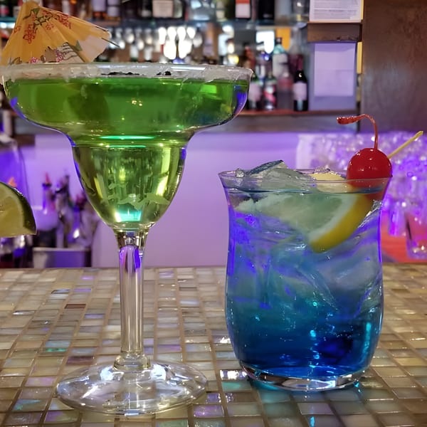 Two vibrant cocktails, one green with an umbrella and one blue with a cherry, on a tiled bar.