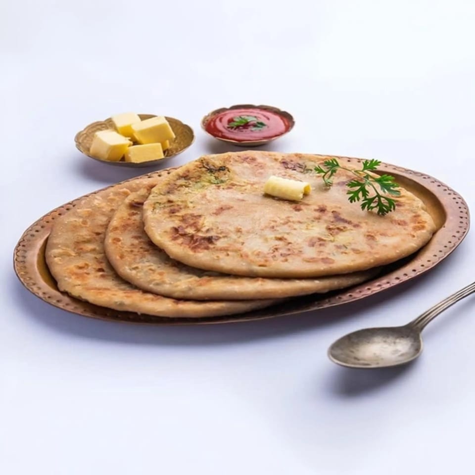 Three Indian parathas on a bronze plate with butter, ketchup, and a spoon.