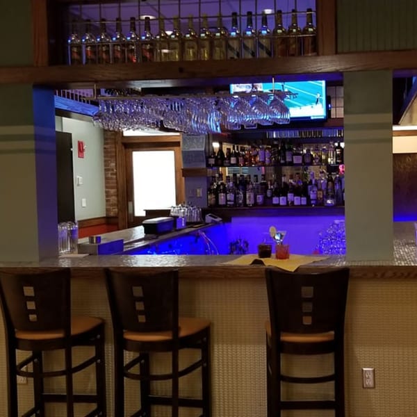 A bar area with blue lighting, bottles on shelves, hanging glasses, and bar stools.
