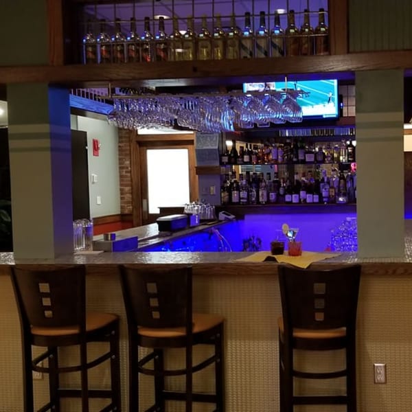 A bar area with blue lighting, bottles on shelves, hanging glasses, and three bar stools.