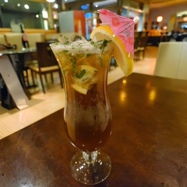 A tall glass of iced tea with lemon slices, mint, and a pink umbrella on a wooden table.