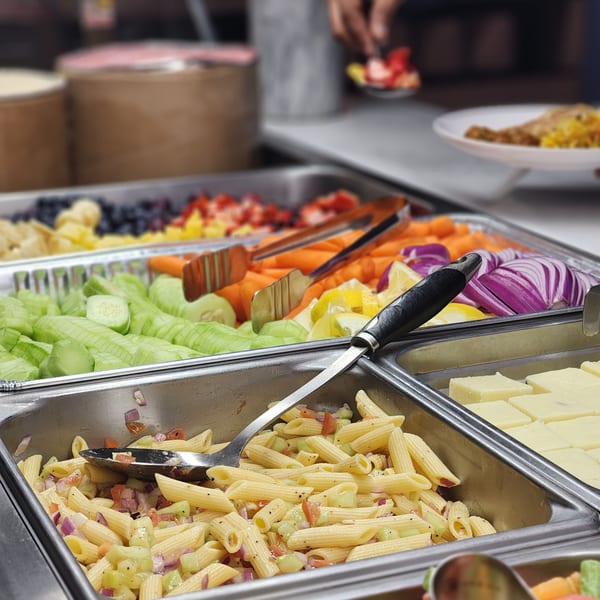 A buffet spread with pasta salad, fresh vegetables, fruit, and Indian sweets.