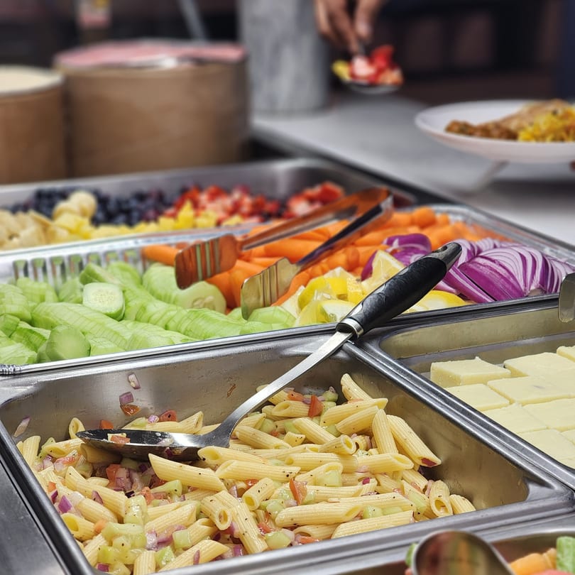 A buffet spread with pasta salad, fresh vegetables, fruit, and Indian sweets.