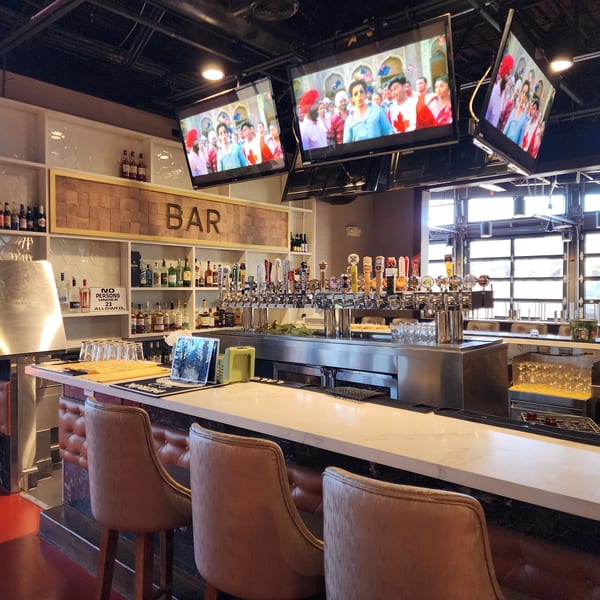 A modern bar area with multiple beer taps, liquor bottles, and TVs showing an Indian movie.
