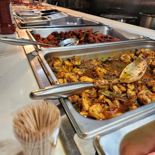 Indian buffet with trays of shrimp curry, red chicken, and other dishes, with serving spoons.