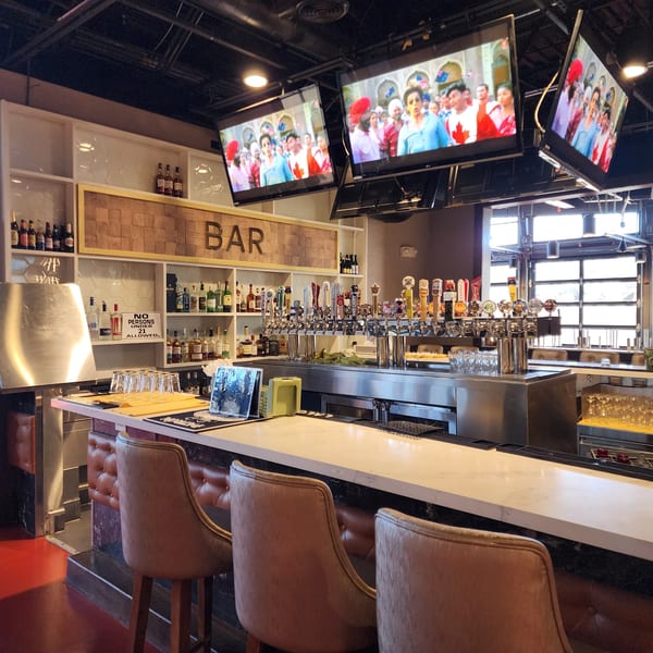 A well-stocked bar at Garnish Restaurants with multiple beer taps, liquor bottles, and TVs showing Indian content.