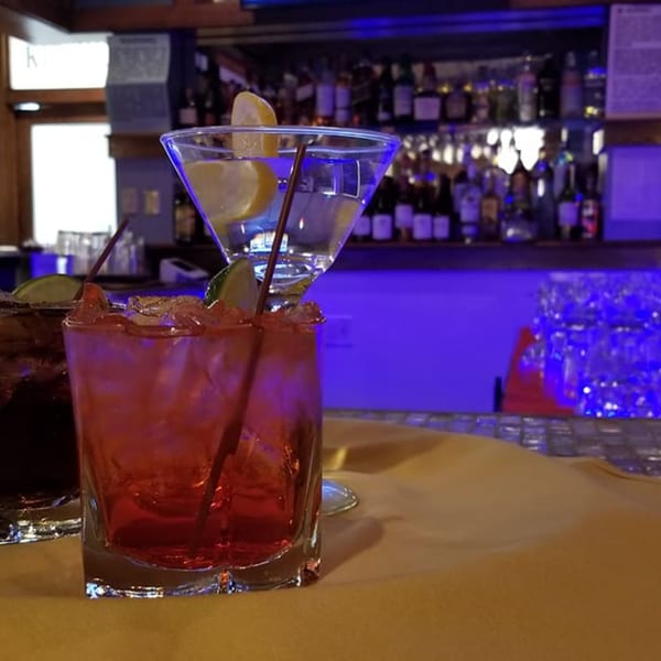 Two cocktails, one red and one clear with lemon, on a table in front of a blue-lit bar.